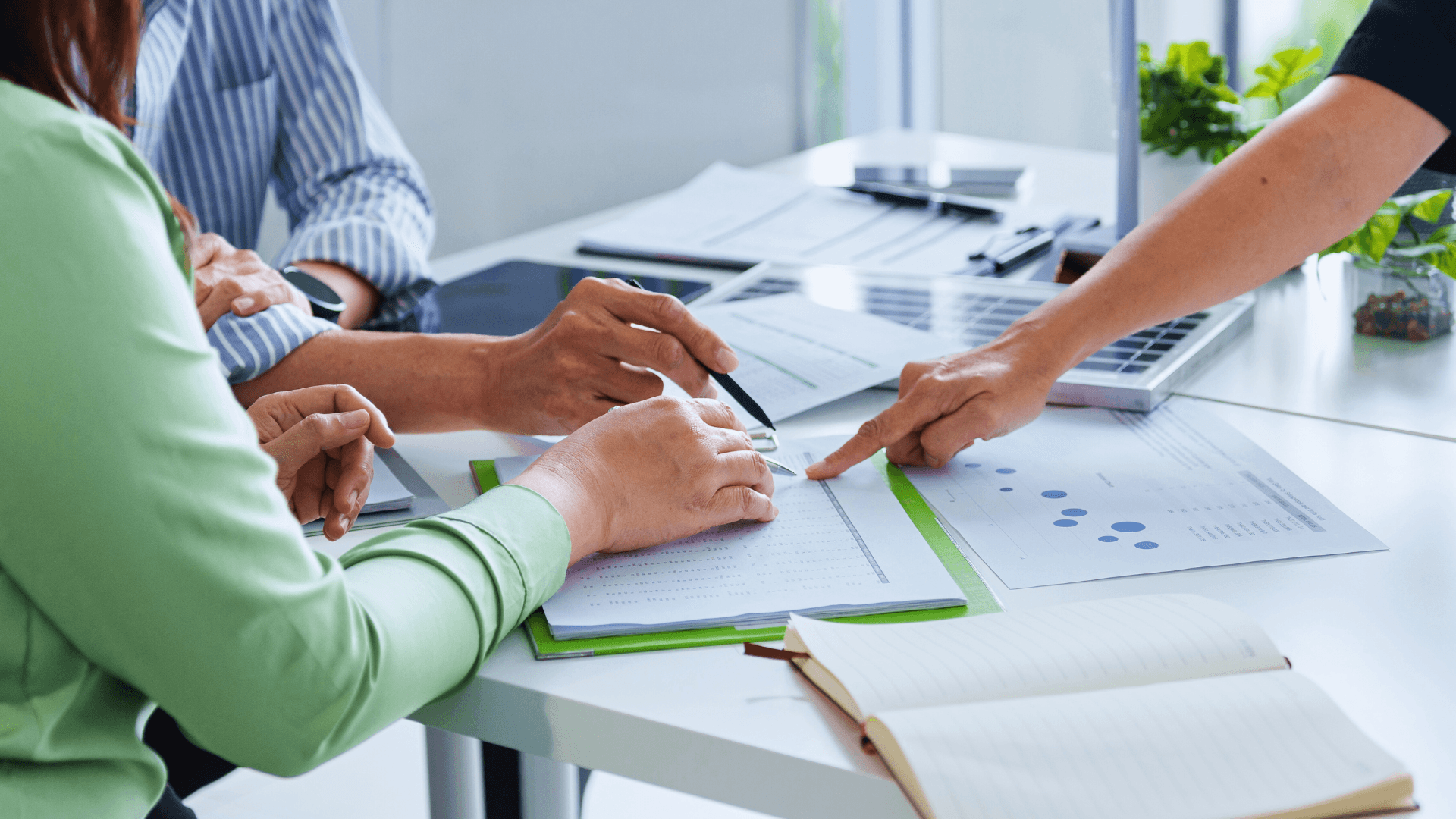 A moment from a business meeting where three people sitting at a table are examining documents and graph paper on a green folder, taking notes with pens.