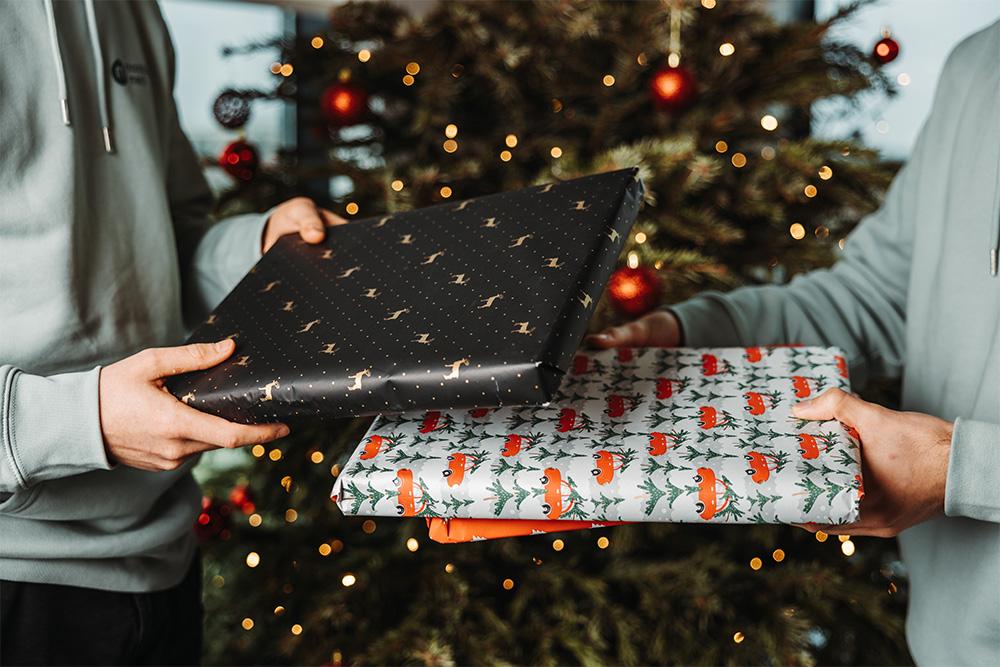 Two people exchanging wrapped gifts, with a Christmas tree in the background