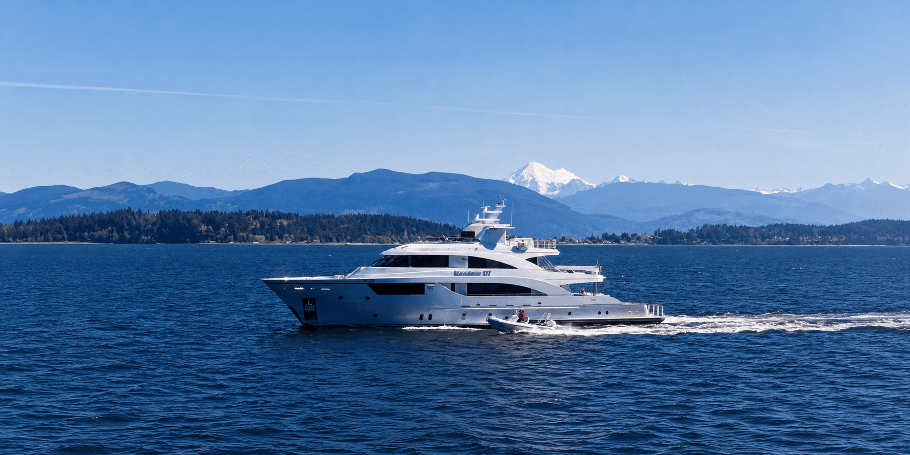 MAXIMUS III cruising across blue water with forested hills and snowcapped mountains in the background.