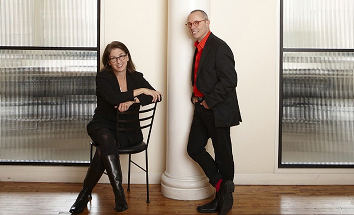 A photo of a woman siting on a chair and a man leaning against a white stone pillar in a room.