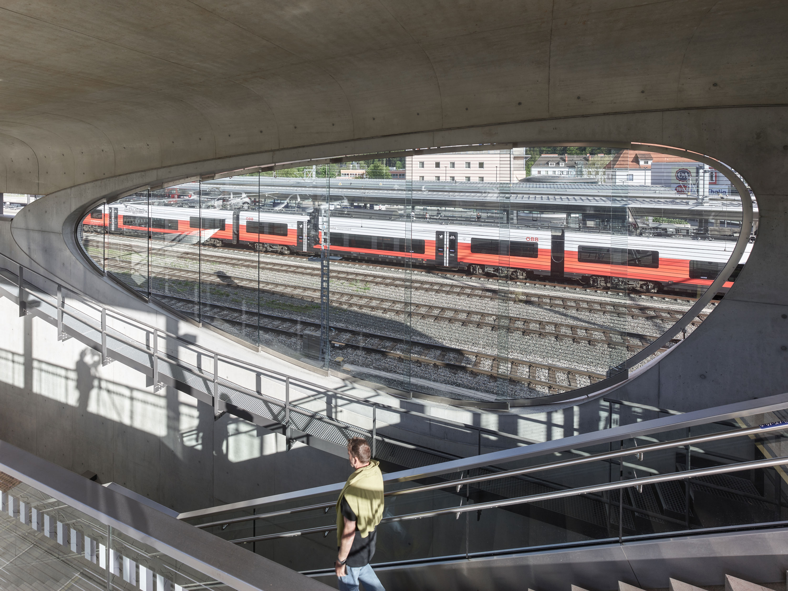 Blick durch ein ovales Fenster im Beton auf Bahngleise mit roten Regionalzügen, Glasfassade, Schattenmuster auf Treppe.