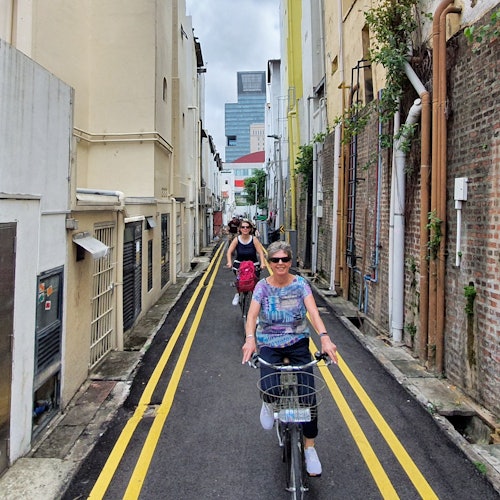 สองคนกำลังปั่นจักรยานลงซอยแคบที่มีอาคาร, ท่อ, และเครื่องหมายถนนสีเหลืองเรียงรายอยู่ สังเหตอาคารสูงได้จากระยะไกล