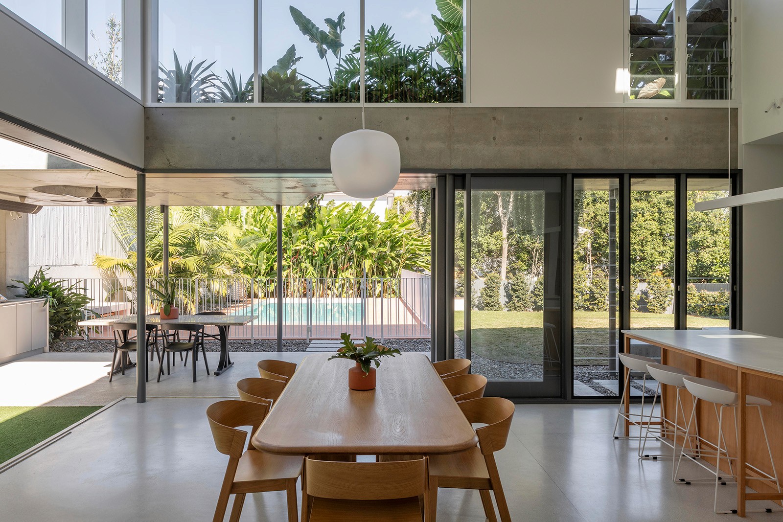 Open-plan dining space with double-height glazing, timber furniture, and seamless views to garden and pool.