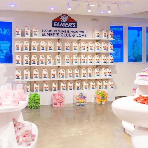 Una exhibición de botellas de pegamento de Elmer organizadas en estantes en una tienda bien iluminada, con varios productos de slime coloridos en estanterías blancas.