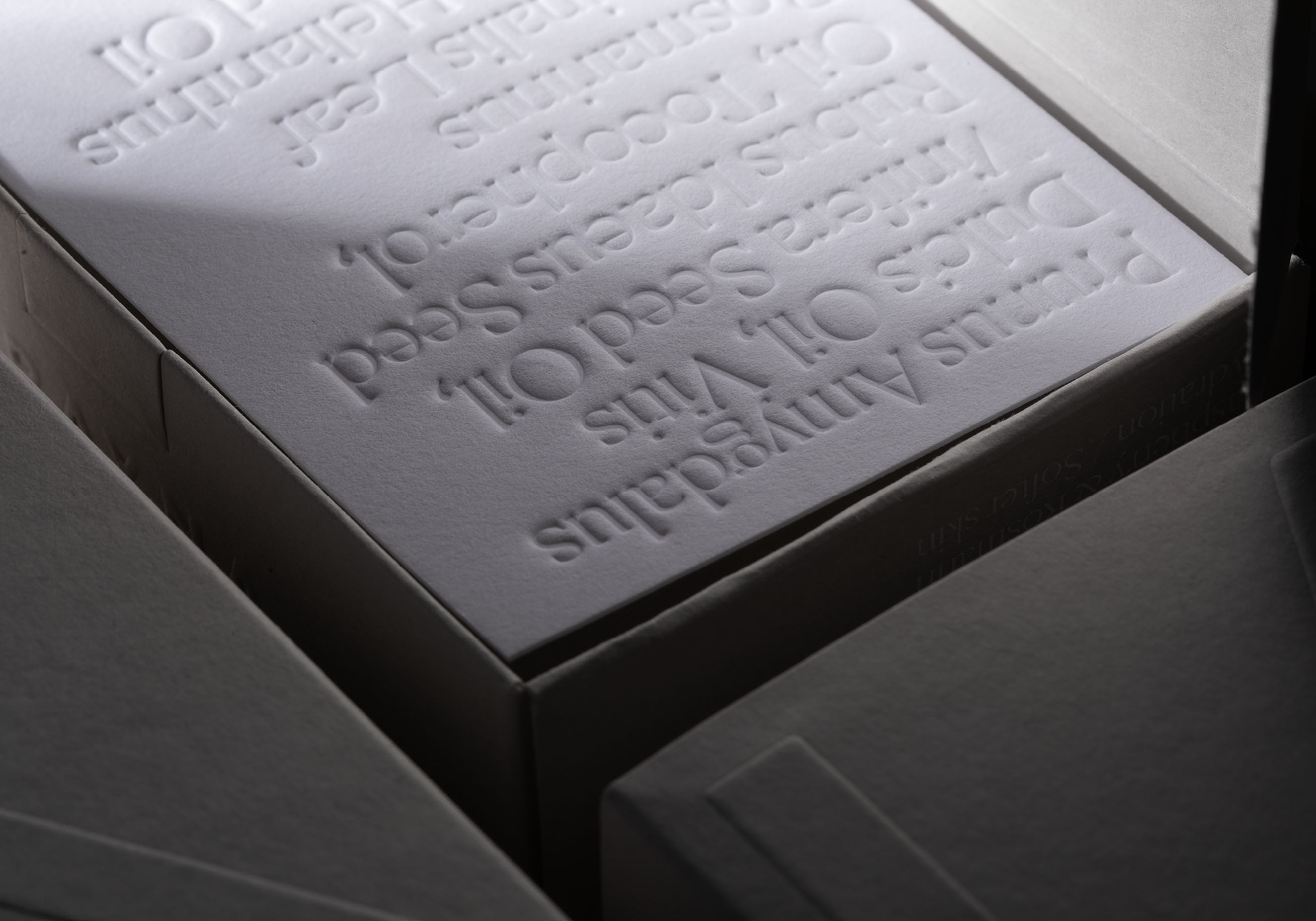 A macro shot of a white paper box featuring deeply embossed botanical names like "Prunus Amygdalus Dulcis Oil." This showcases a clean, information-heavy design that maintains a luxury feel for a transparent, vegan brand.