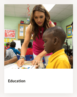 Teacher leans over to help a young student at a desk in a colorful classroom. Other students are visible in the background. The word “Education” appears below the image.