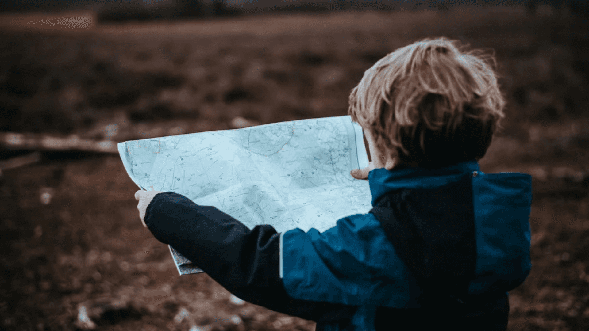 A child seen from behind holding and studying a folded paper map while standing in an open, brown-toned landscape.