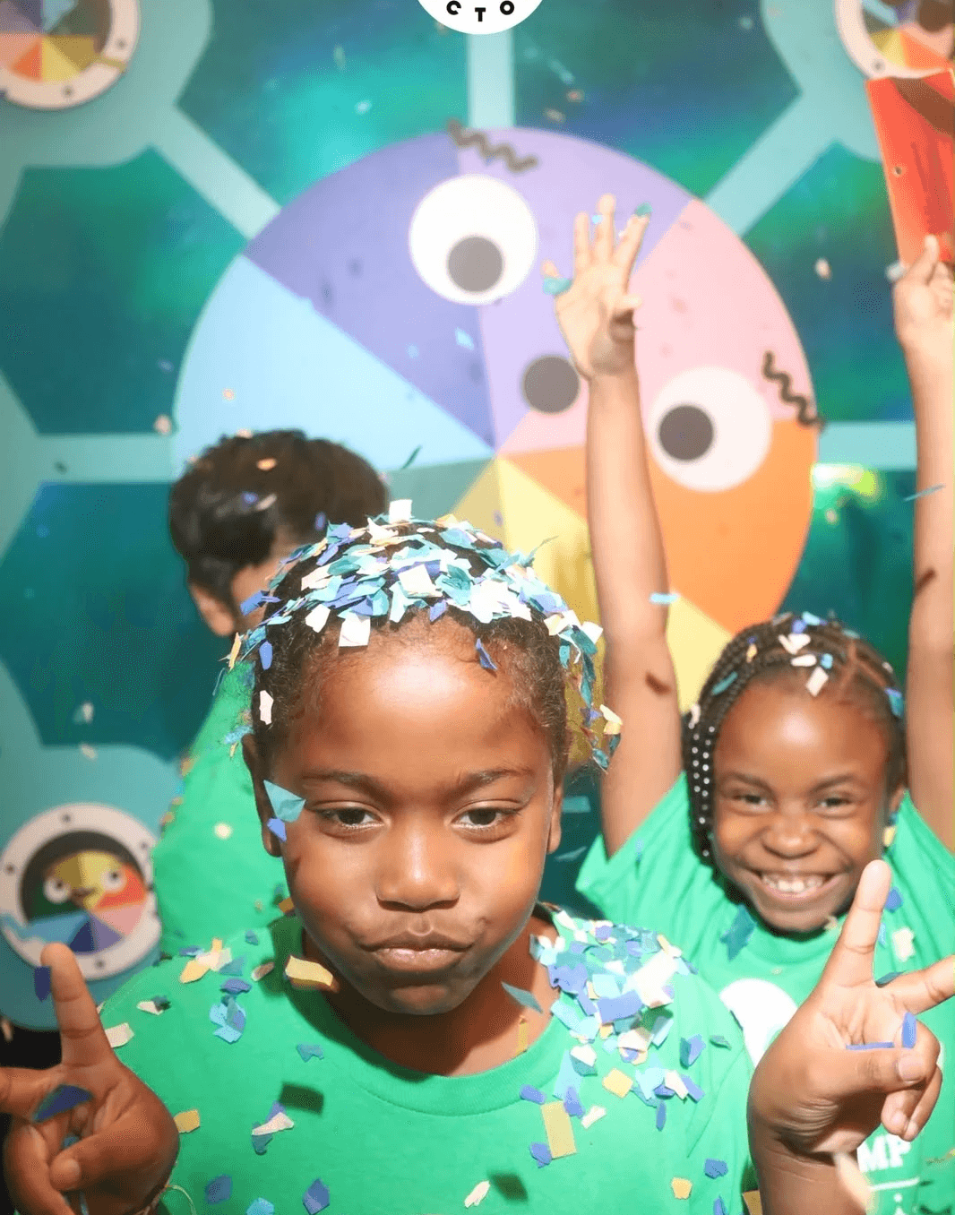 Campers covered in colorful confetti pose in front of a colorful wheel with googly eyes.