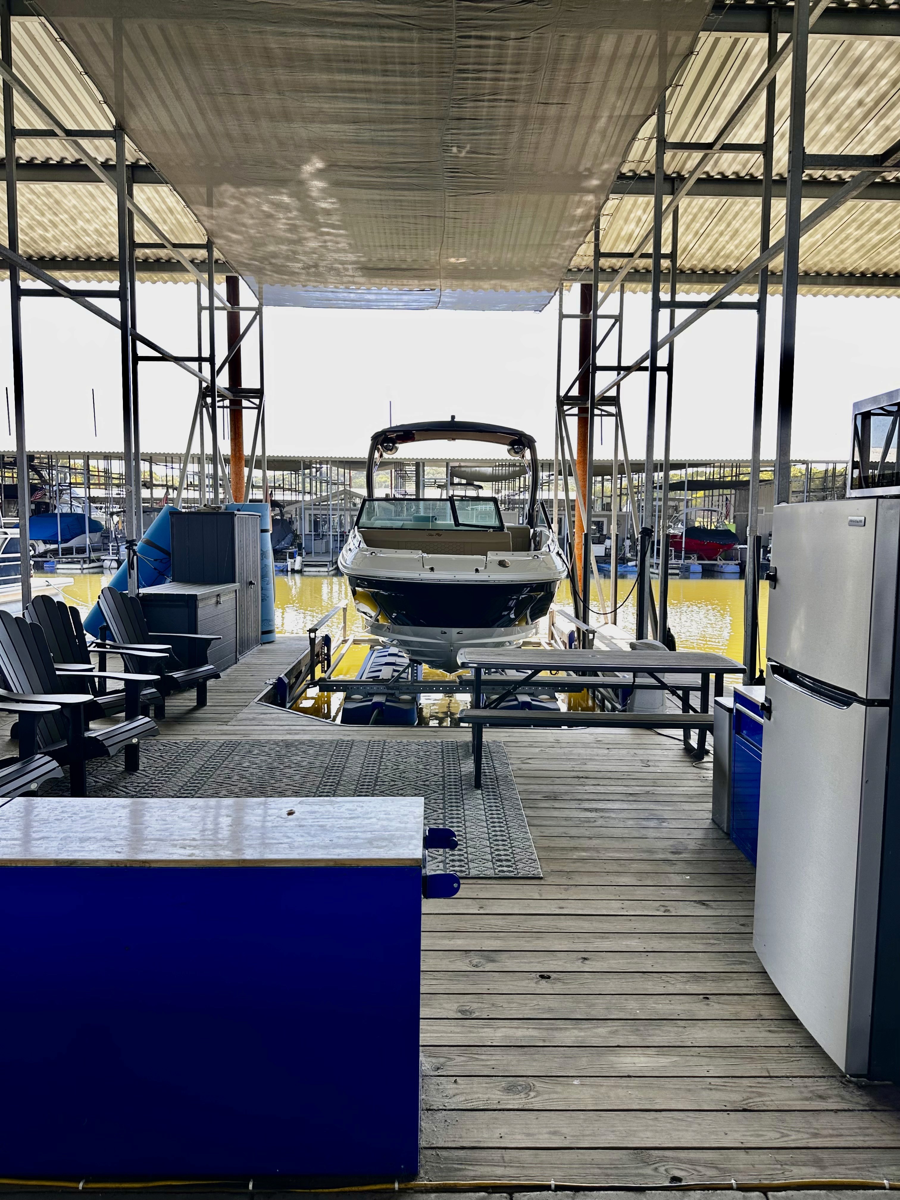 A motorboat is docked inside a covered marina, surrounded by outdoor seating, a refrigerator, and storage containers, with calm water visible in the background.