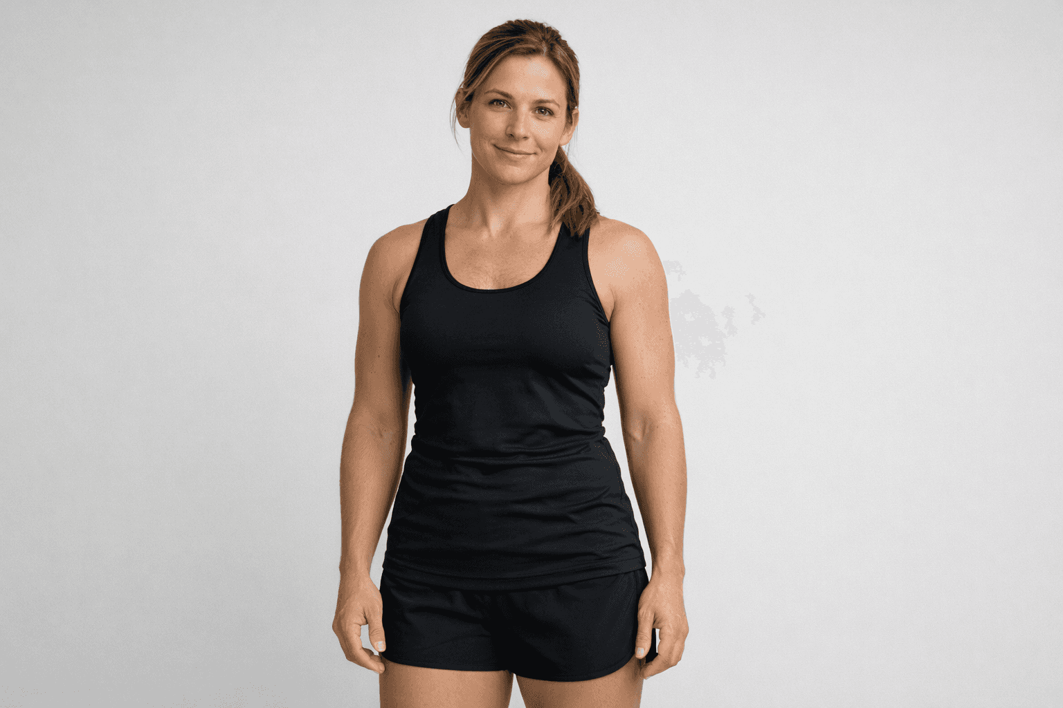 Fit woman in black athletic tank top and shorts smiling against a neutral studio background.
