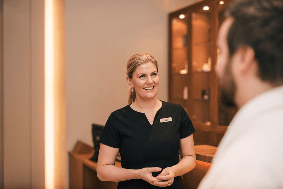 A friendly staff member in a black uniform stands smiling and interacting with a customer in a warmly lit hotel reception area.