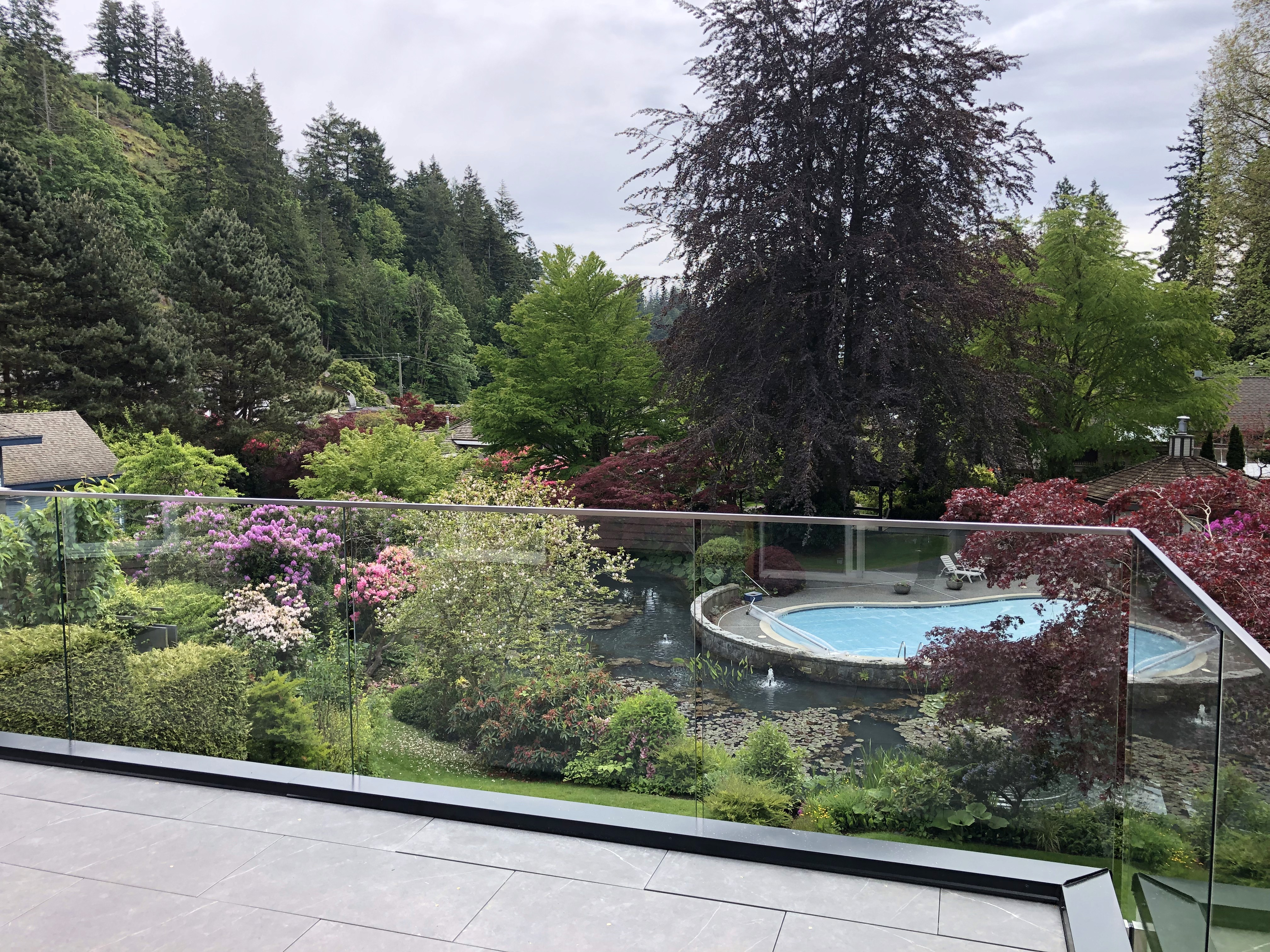 Frameless 12mm glass railing on a Pacific Northwest deck overlooking a landscaped garden, pool, and forested hillside