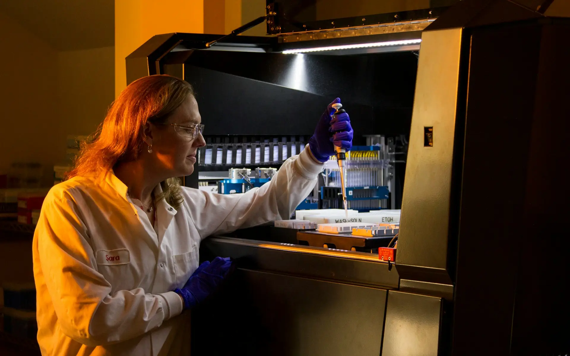 Scientist in a lab coat using a pipette in a dimly lit laboratory with advanced equipment in the background.