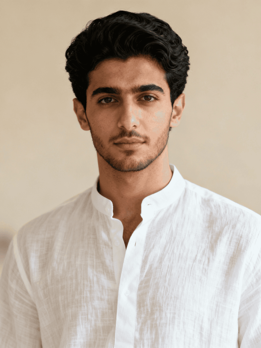 Portrait of a young man with dark wavy hair wearing a white linen shirt against a warm beige studio background.