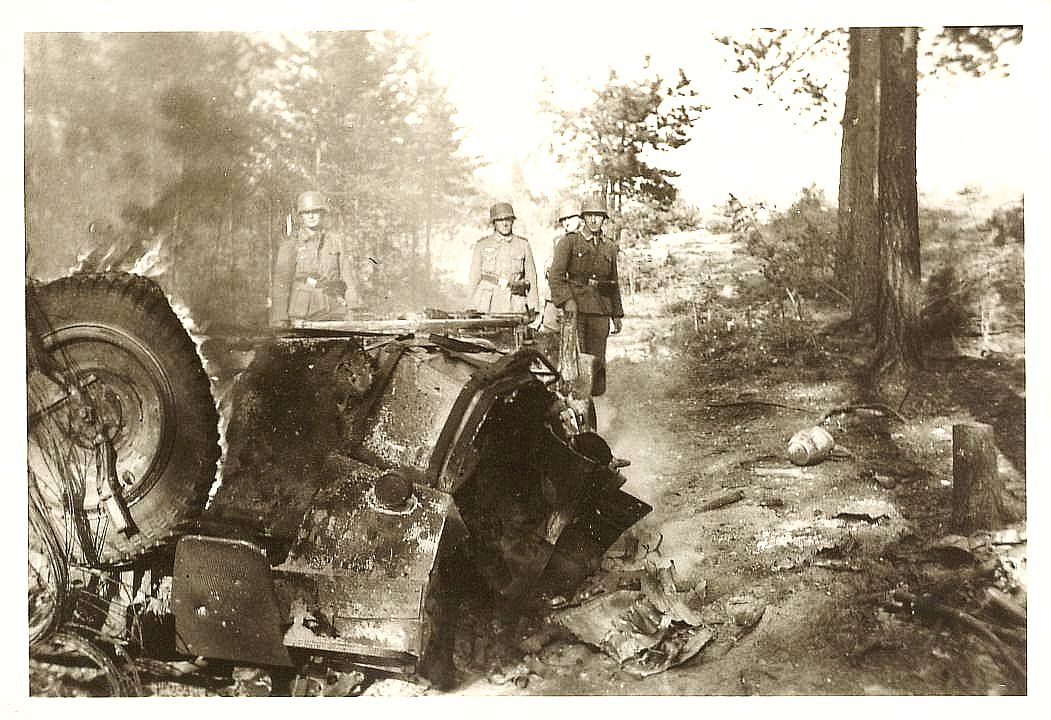 Fire tyske soldater står ved vraget af et ødelagt køretøj i en skov under Anden Verdenskrig. Billedet viser det ødelagte landskab, med brand og røg stige op fra vraget og træer i baggrunden.