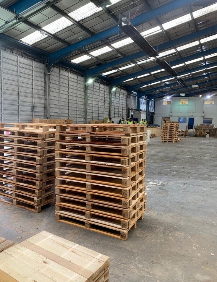 Stacks of wooden pallets in a spacious warehouse with a high ceiling. Workers in safety vests are gathered in the background, creating a busy industrial atmosphere.