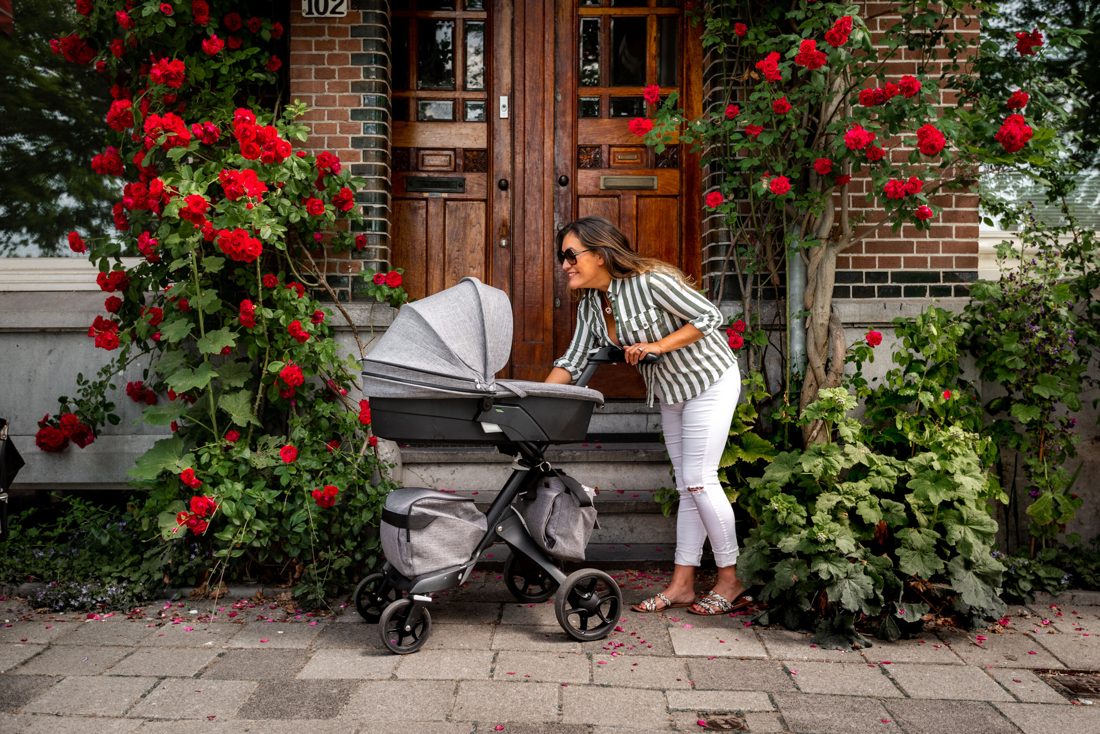 Sunlight illuminates a woman beside a stroller on a brick walkway lined with vibrant red roses and greenery.