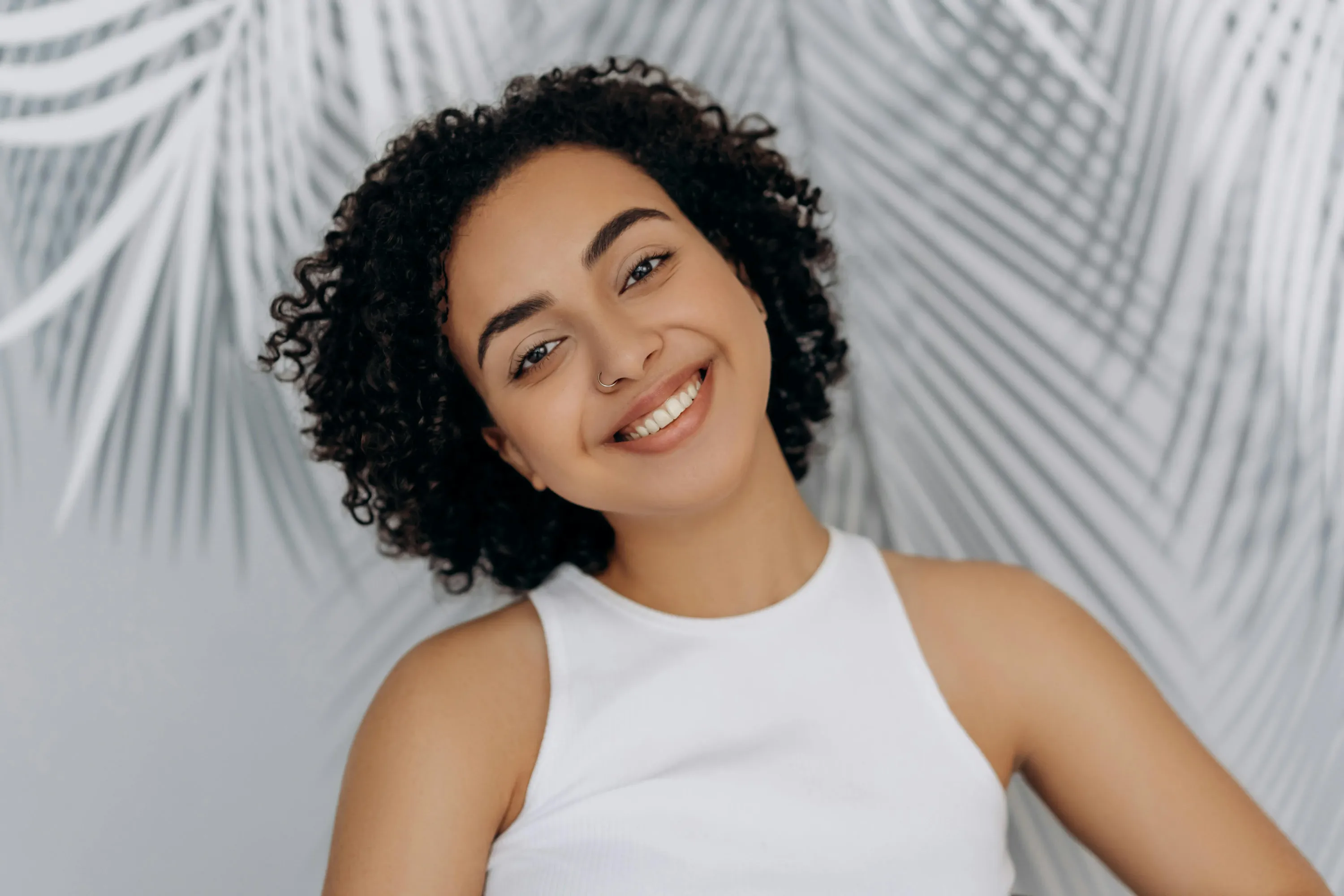 A warm and inviting close-up portrait of a smiling woman with voluminous curly hair and a white tank top, posed naturally in front of soft grey palm leaf patterns.