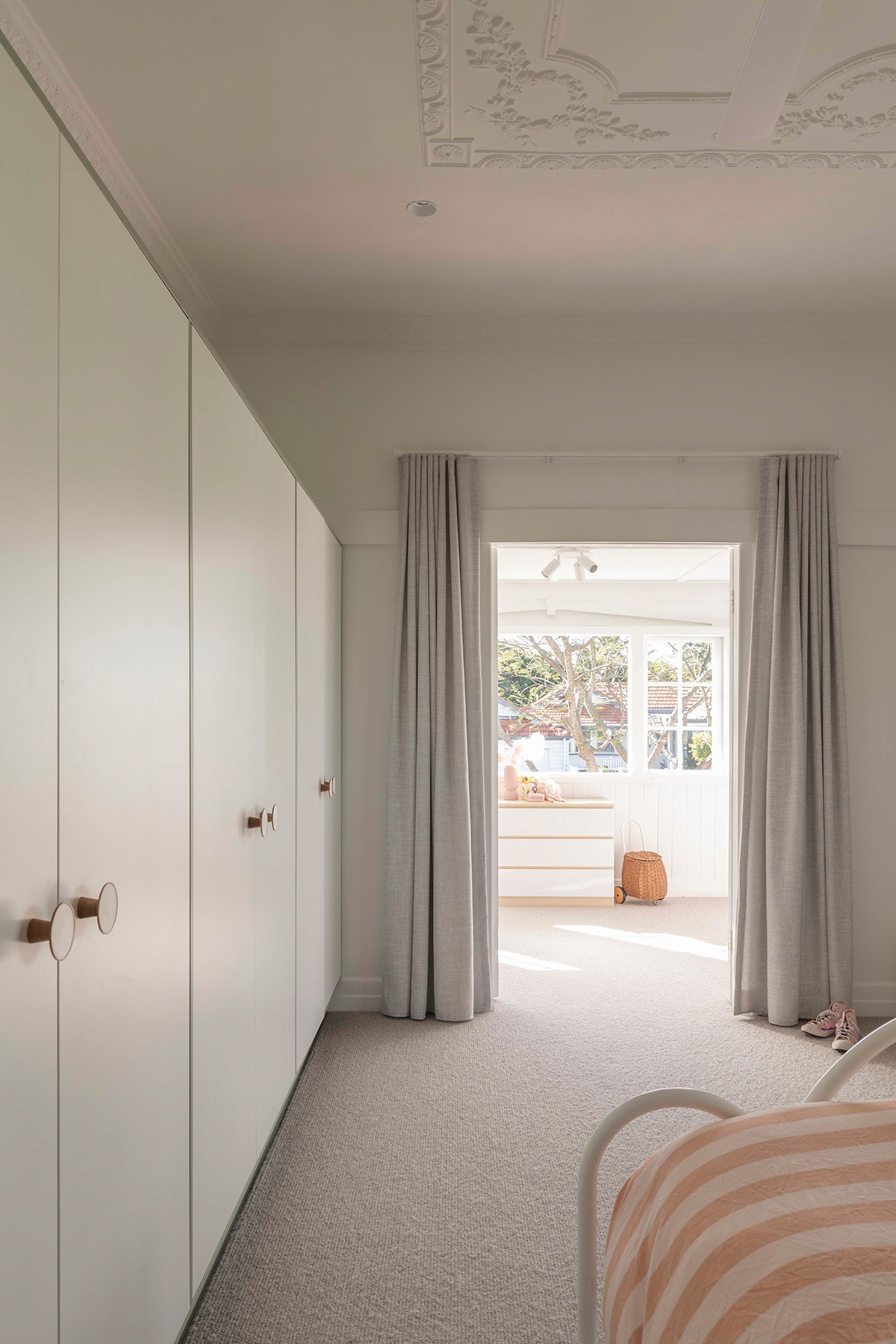Light-filled bedroom with built-in wardrobes, soft drapery, and decorative ceiling detail opening through to a sunlit adjoining space.