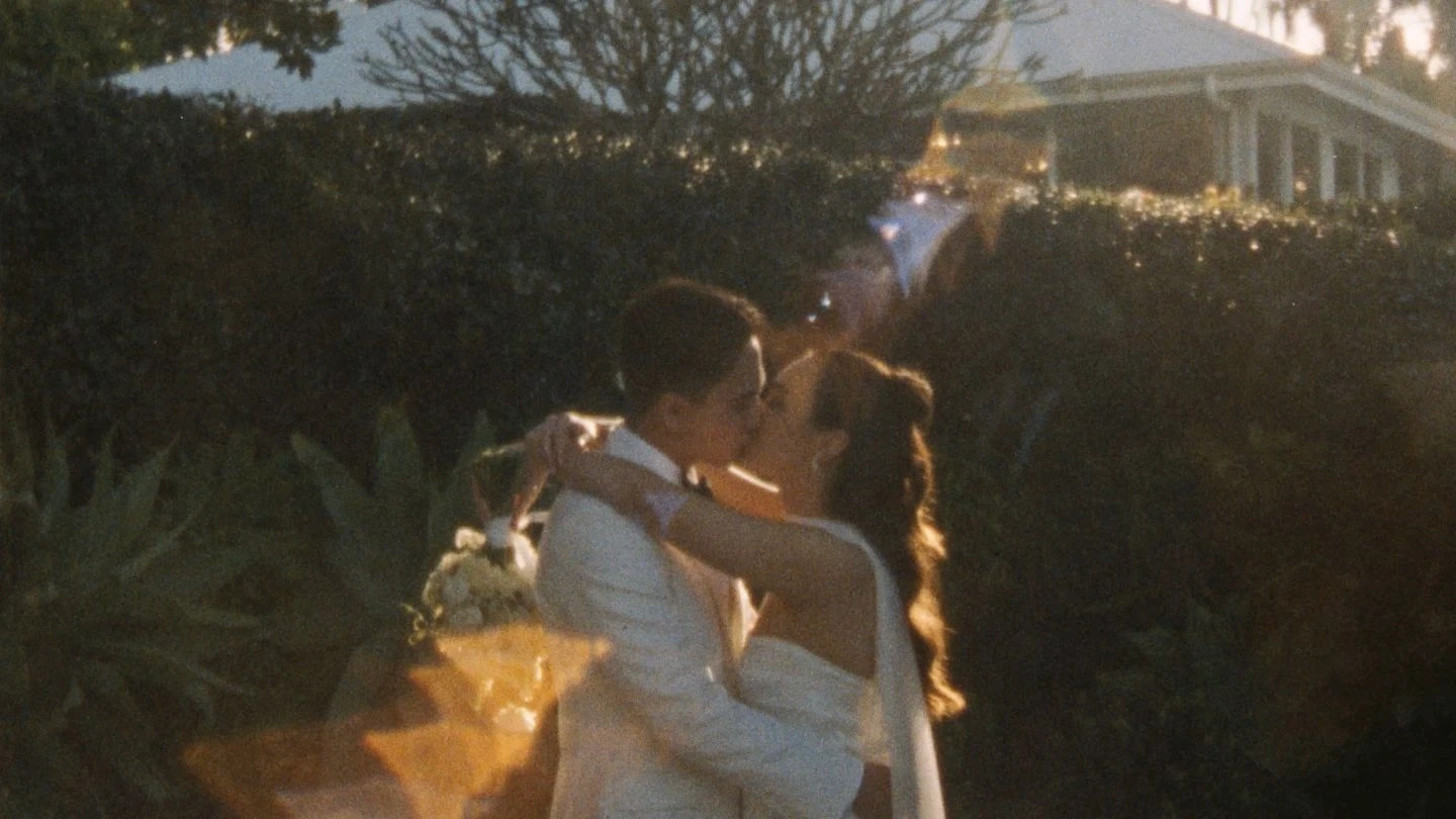 A couple shares a romantic kiss in a garden setting, with the sunlight creating a warm glow around them; the groom is dressed in a white suit, while the bride holds a floral bouquet, adding to the enchanting, love-filled atmosphere.