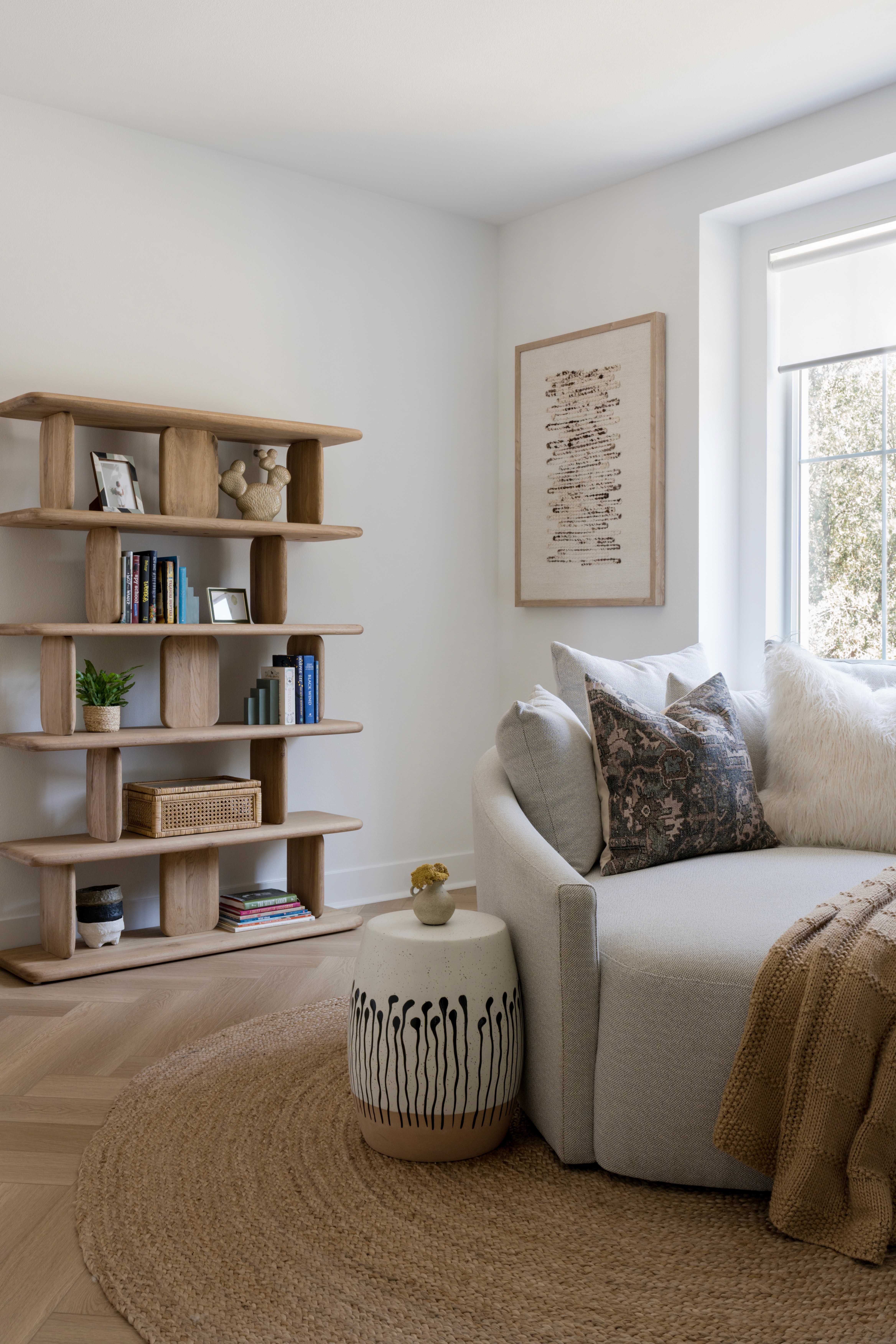 Loft with neutral wood book shelf, off-white love seat, woven rug, mustard throw blanket, ceramic side table, light and dark decorative pillows, and modern wall art by Jubilee Interiors in Arcadia, California