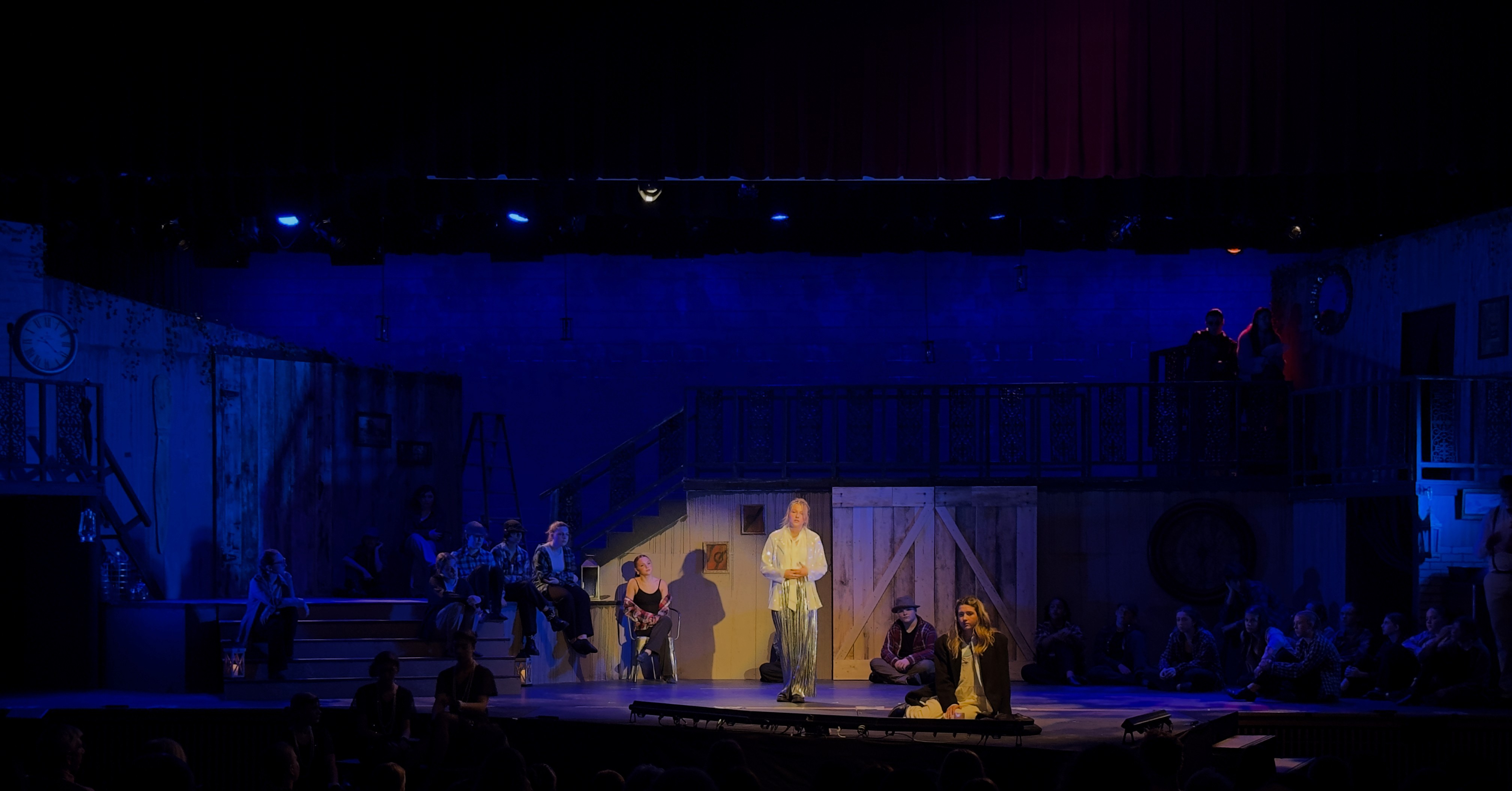 Any Way the Wind Blows, a scene from Hadestown, is set under a cool blue stage wash, with somber lighting creating a dramatic and moody atmosphere, featuring actors performing in front of a rustic, darkened background.