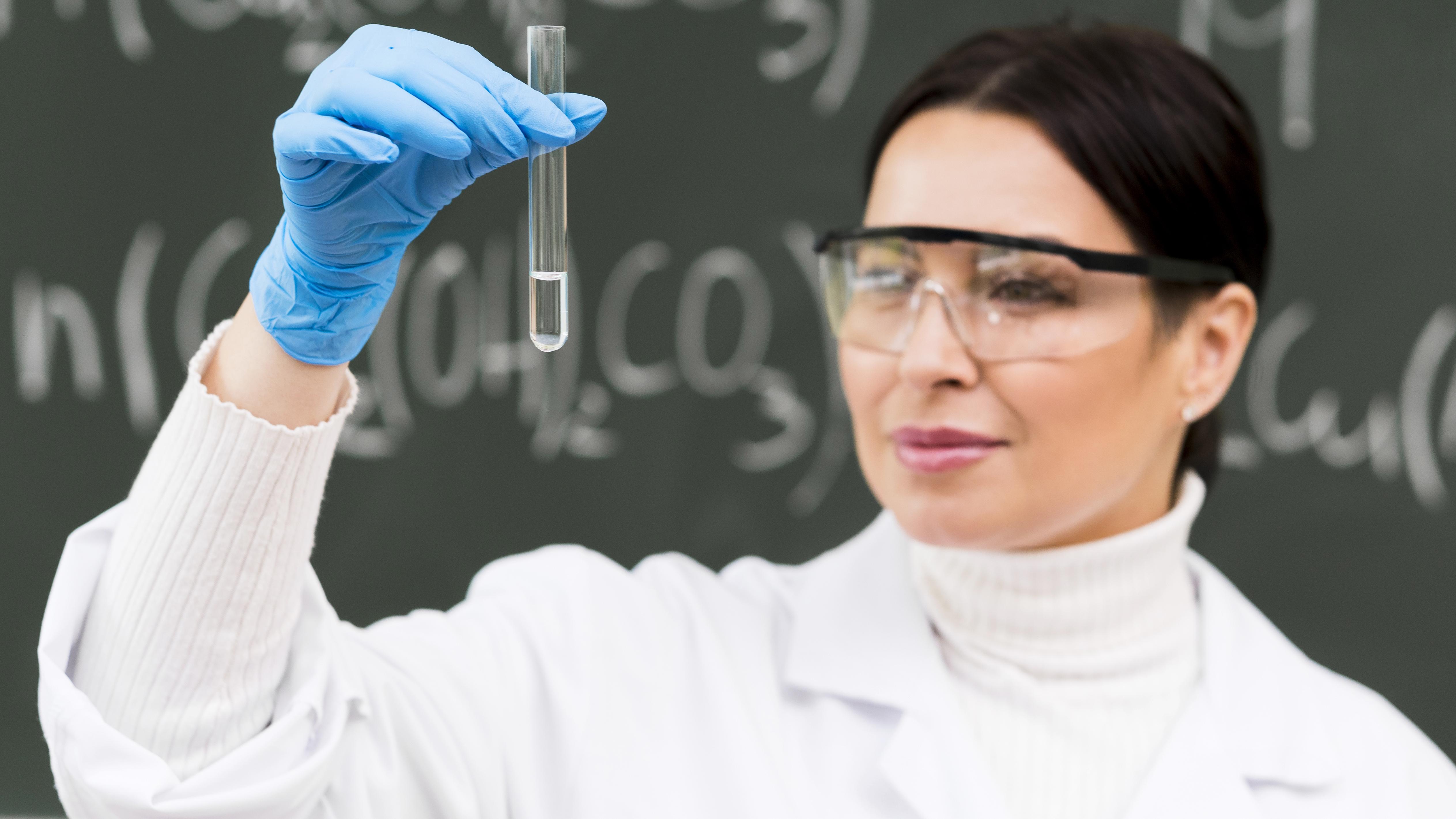Female scientist in a lab wearing safety goggles and holding a test tube against a background of chemical formulas.