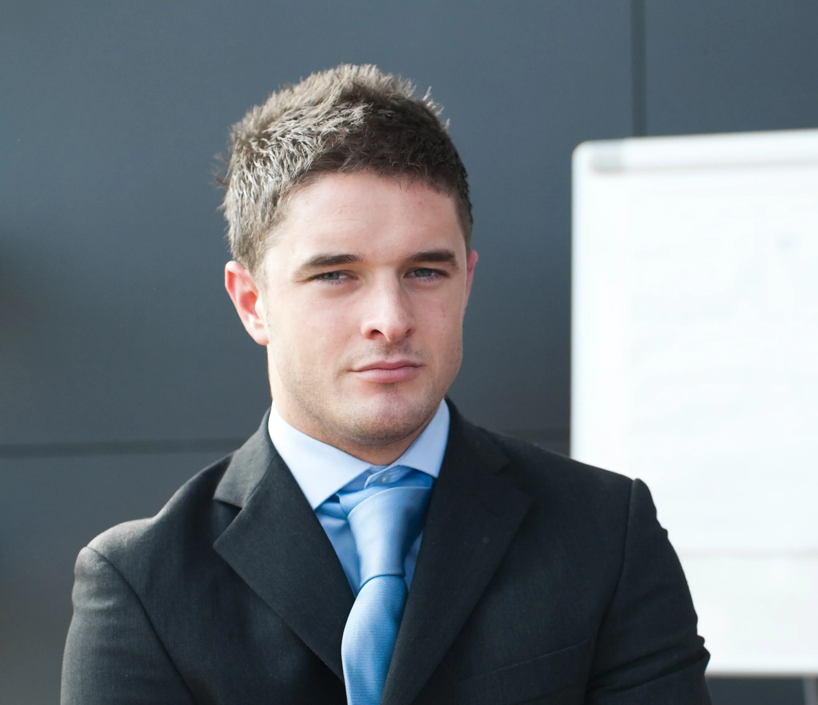 Young professional male in a black blazer and light blue tie looking forward in a clean office setting