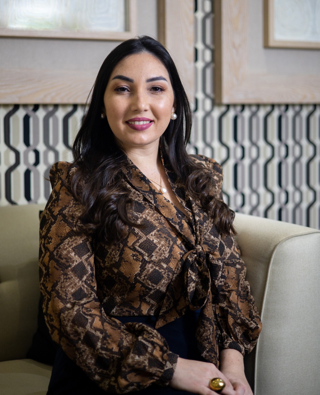 Psicóloga Vera Joubert sentada en un sofá color crema, posando profesionalmente, con una blusa estampada en tonos marrones y cuello de lazo.