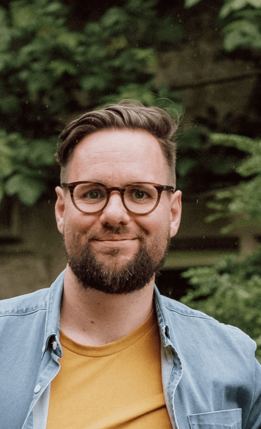 A smiling person with curly hair sits at a desk, looking at a laptop against a dark background.