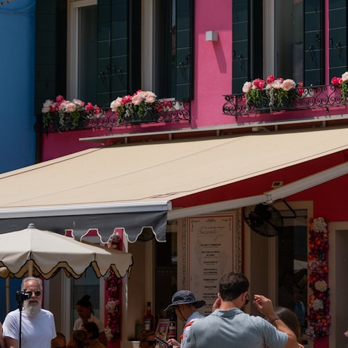 一处街景,粉色建筑的阳台饰有鲜花,设有遮阳篷,下方聚集着人群。