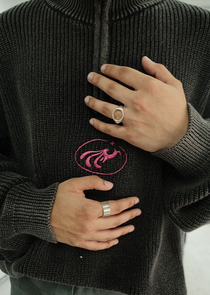 Person wearing a dark sweater and silver jewelry with a pink logo, holding their hands close to the chest.