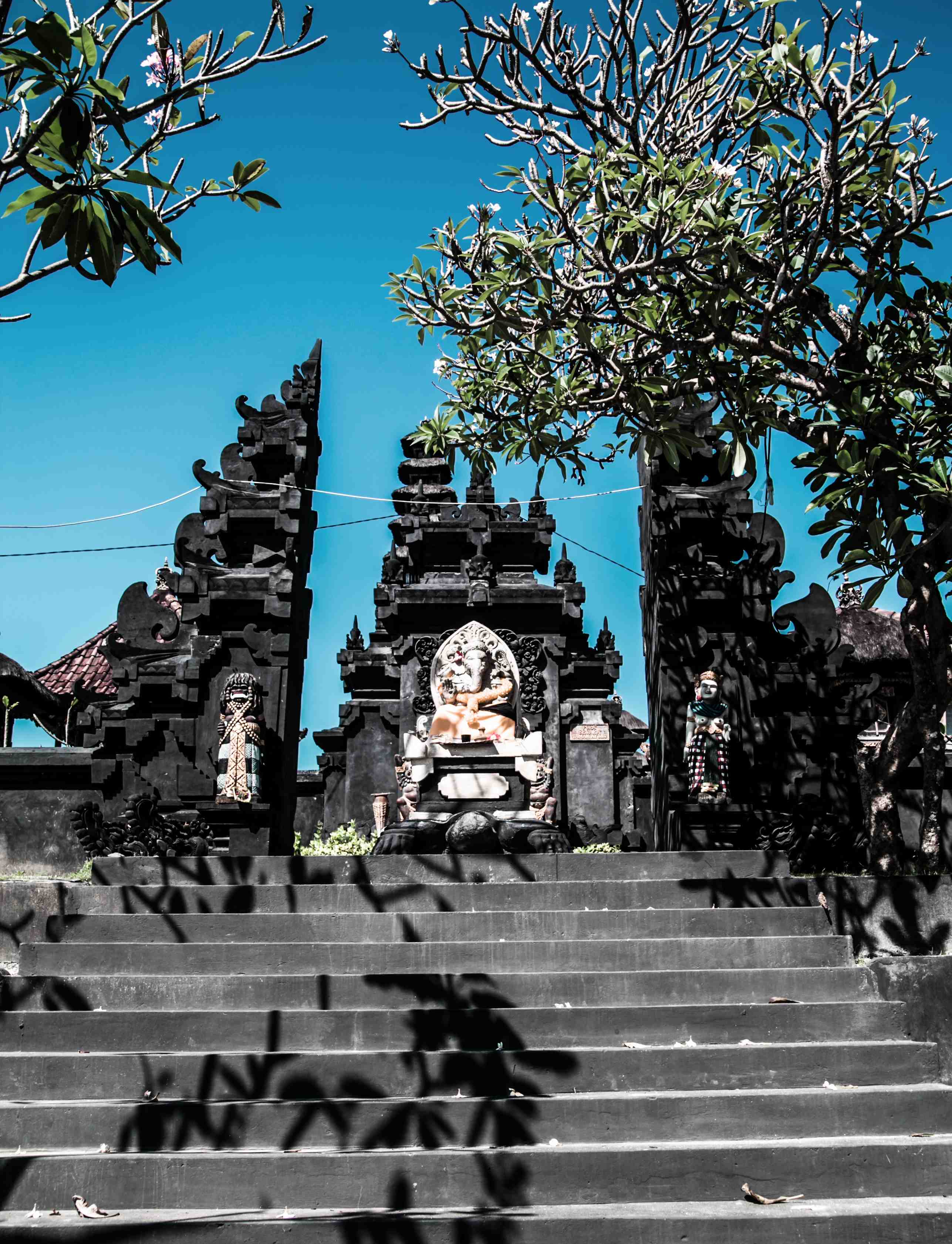 Steps lead to an intricately carved, dark stone Balinese temple gate under a clear blue sky in Ubud. Framing trees cast dynamic shadows, evoking serenity.