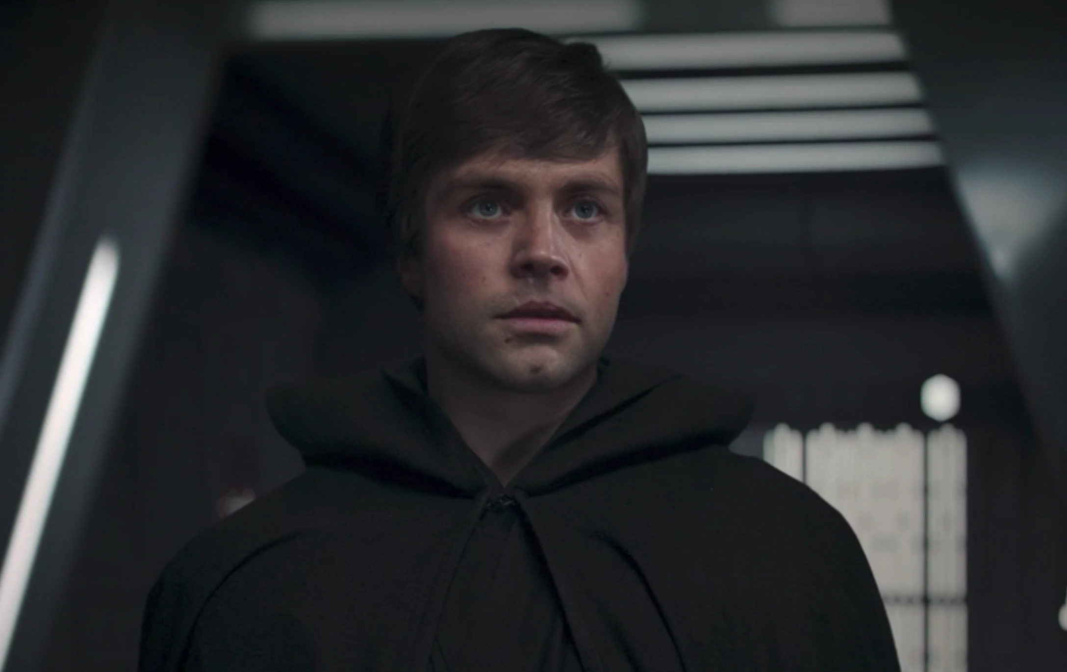 Luke in a dark cloak stands in a dimly lit corridor, looking thoughtful or concerned.