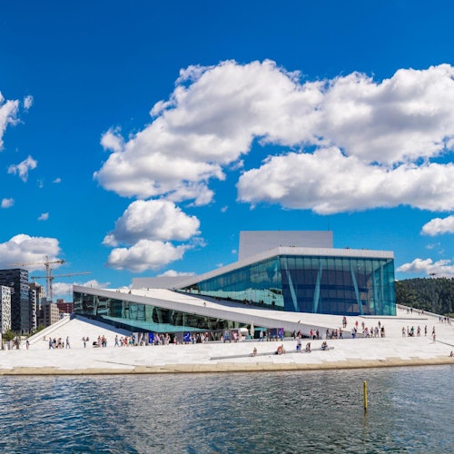 Modern building with a sloped roof, people walking, bright blue sky with clouds, and water in the foreground.
