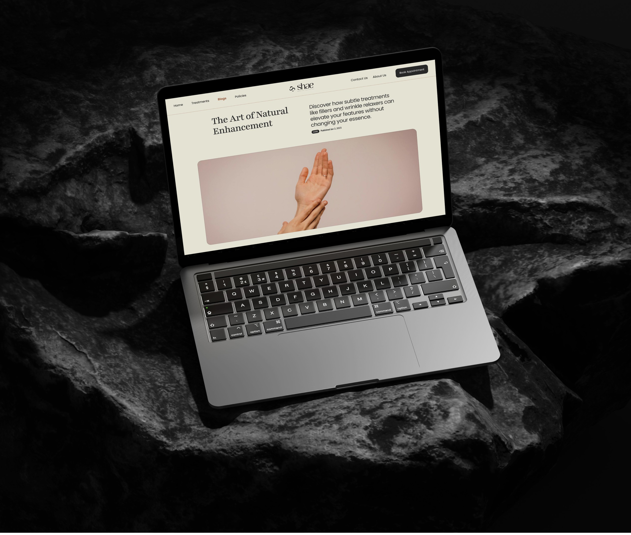 Laptop displaying the Shae Medical Aesthetics website with a section titled 'The Art of Natural Enhancement,' featuring an image of hands and text about subtle treatments, placed on a dark, rocky background.