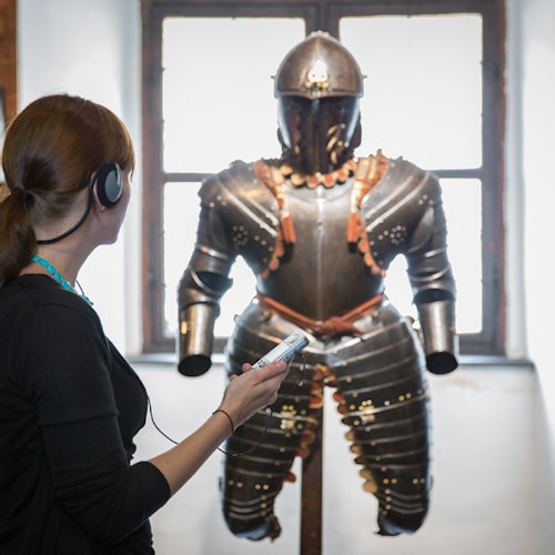 一位戴着耳机的女士正在用语音导览欣赏陈列在大窗户前的盔甲。