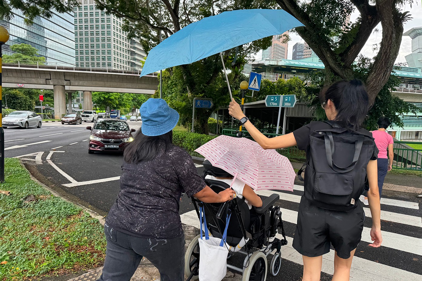 Two caregivers and a wheelchair user outdoors in the rain, with one caregiver holding an umbrella over the other, who is shielding the wheelchair user with a second umbrella