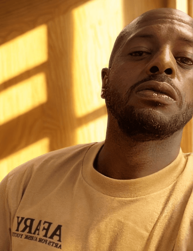 Confident man standing in a greenhouse surrounded by lush green plants, wearing a beige shirt and neutral-toned outfit.