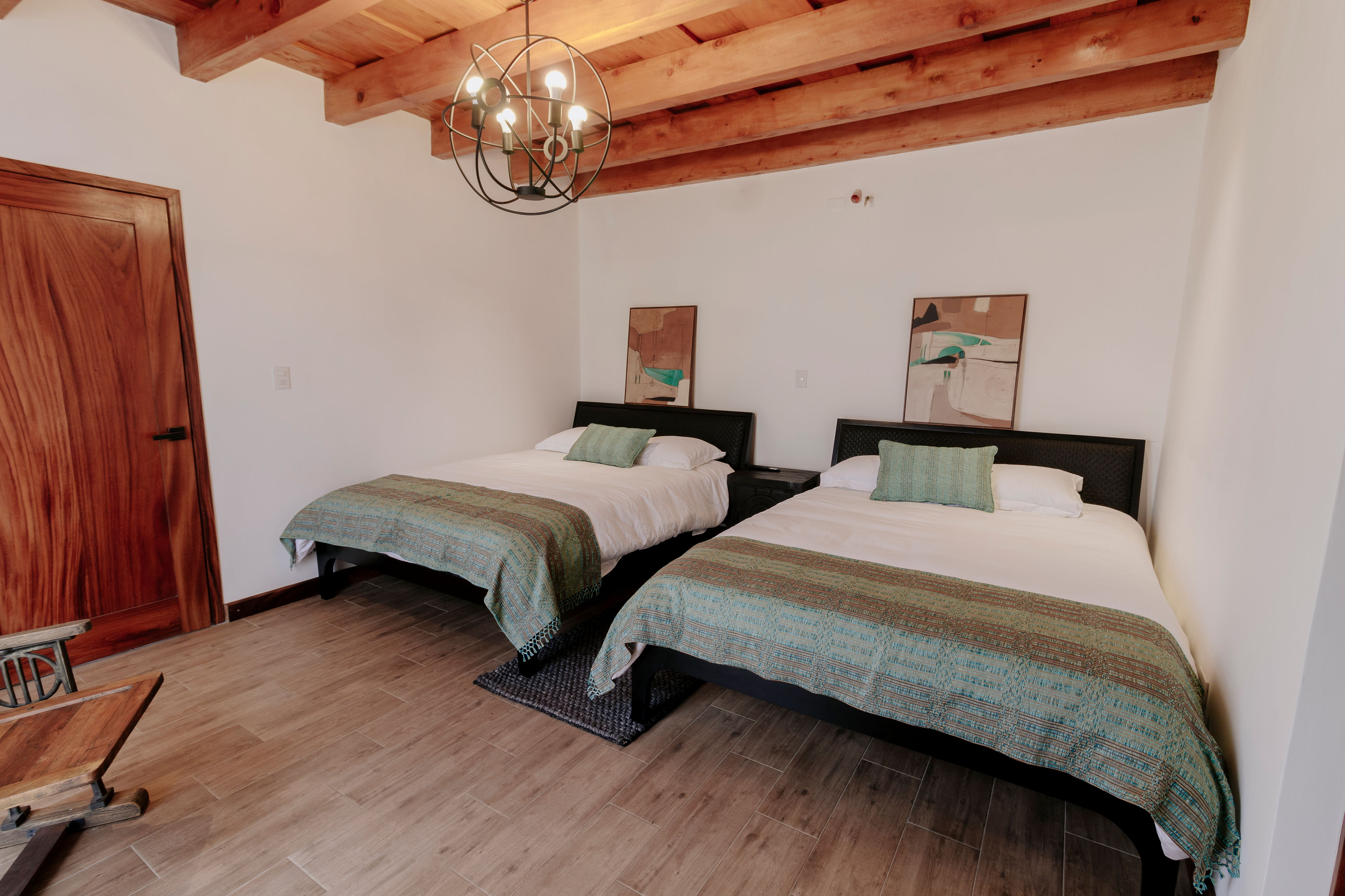 A cozy rustic bedroom features two neatly made double beds with green accent pillows, set against a white wall adorned with abstract art pieces; the room is enhanced by an exposed wooden beam ceiling, a modern chandelier, and a wooden door, creating a warm and inviting atmosphere.