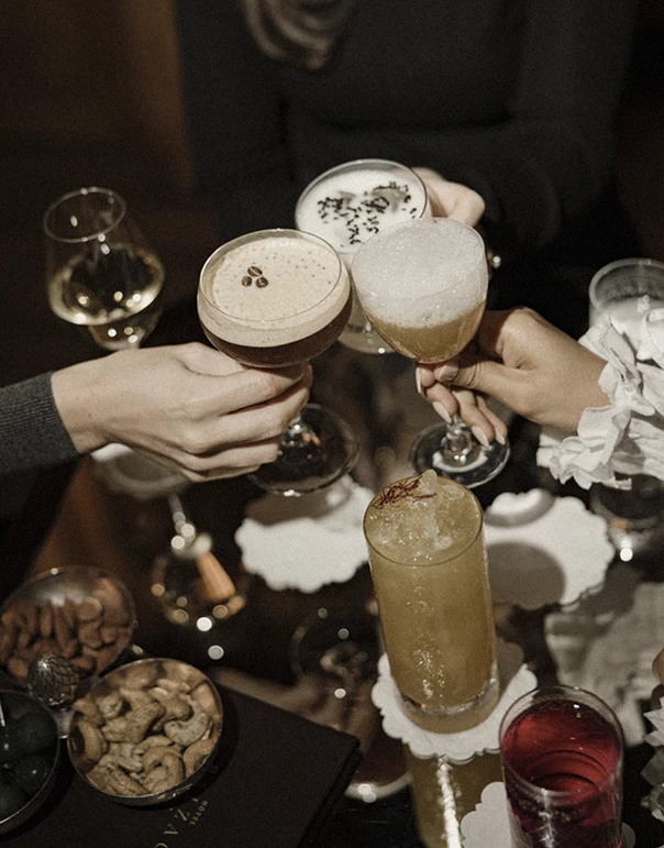 Friends toasting with cocktails in a warmly lit setting, the table filled with drinks and snacks — radiating sophistication and conviviality.  