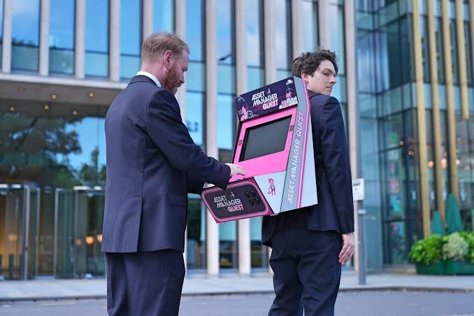 photo of a a man wearing a screen on his back, featuring a video game that another man is playing with. They're both wearing suits and standing in front of a corporate office.