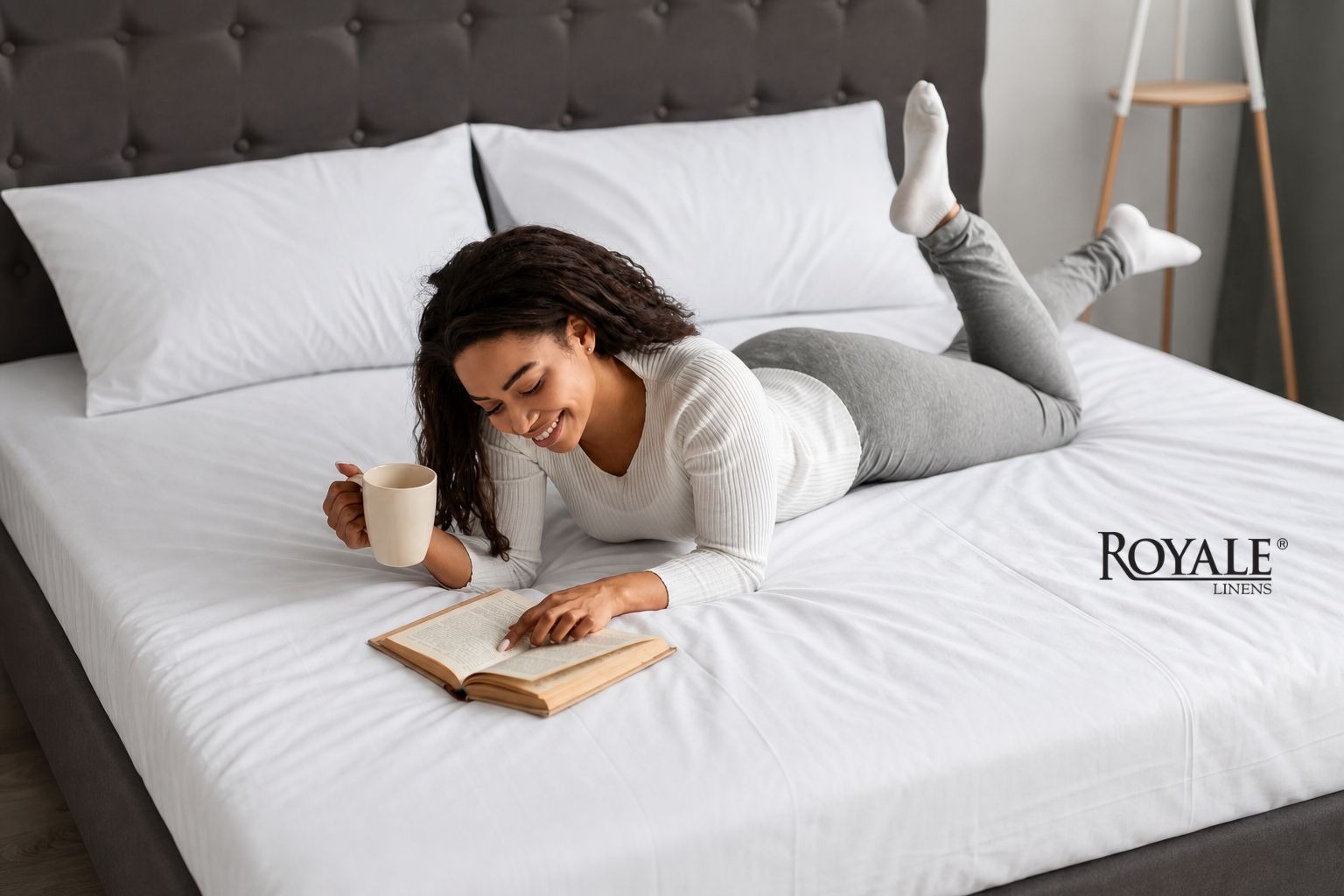 Person relaxing on a bed with Royale Linens bedding while reading a book and holding a mug.