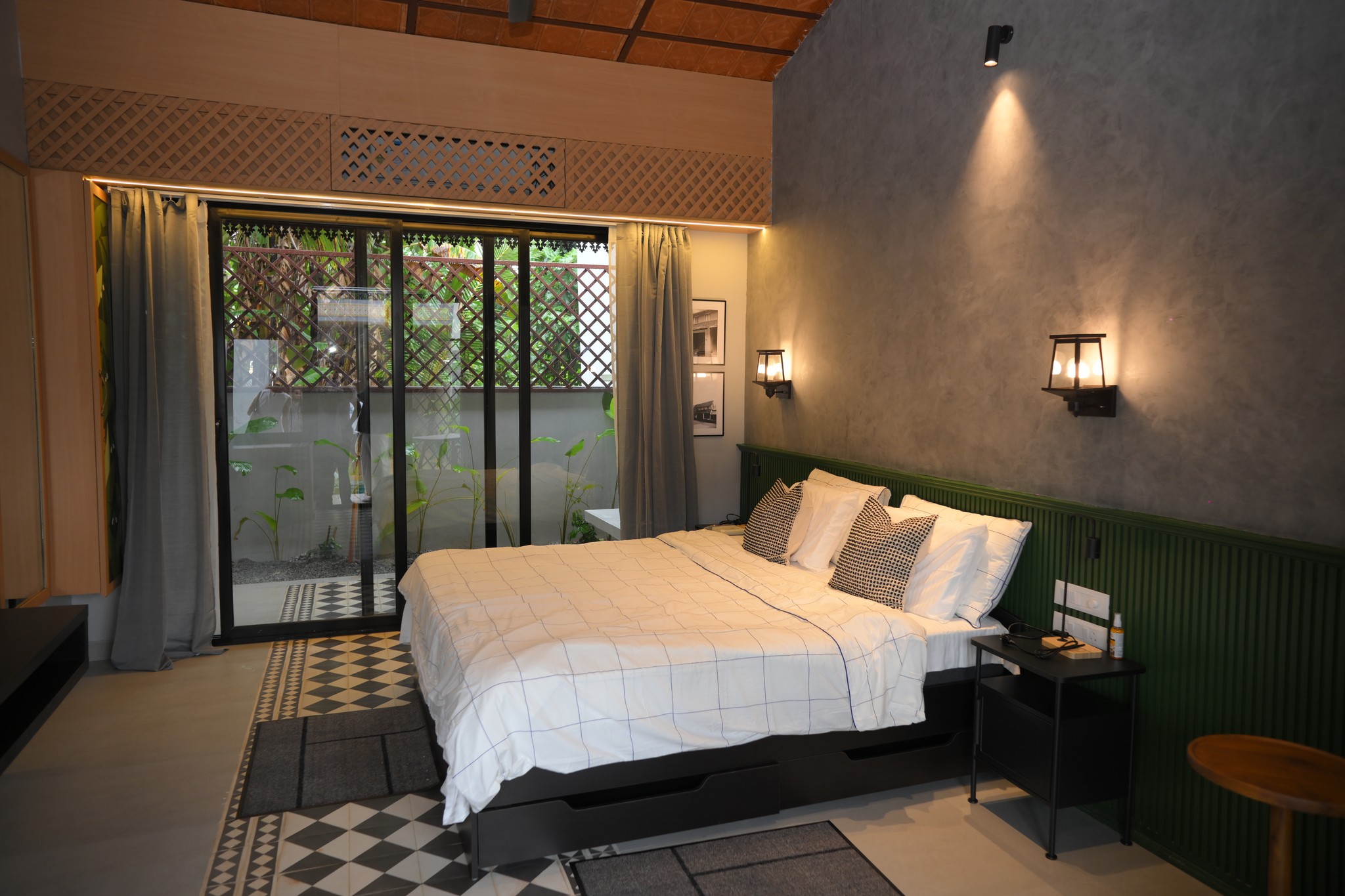 A cozy and inviting spa room with a neatly made bed, soft lighting, and warm wooden tones. The scene is partially framed by green plants, adding a natural and soothing touch.