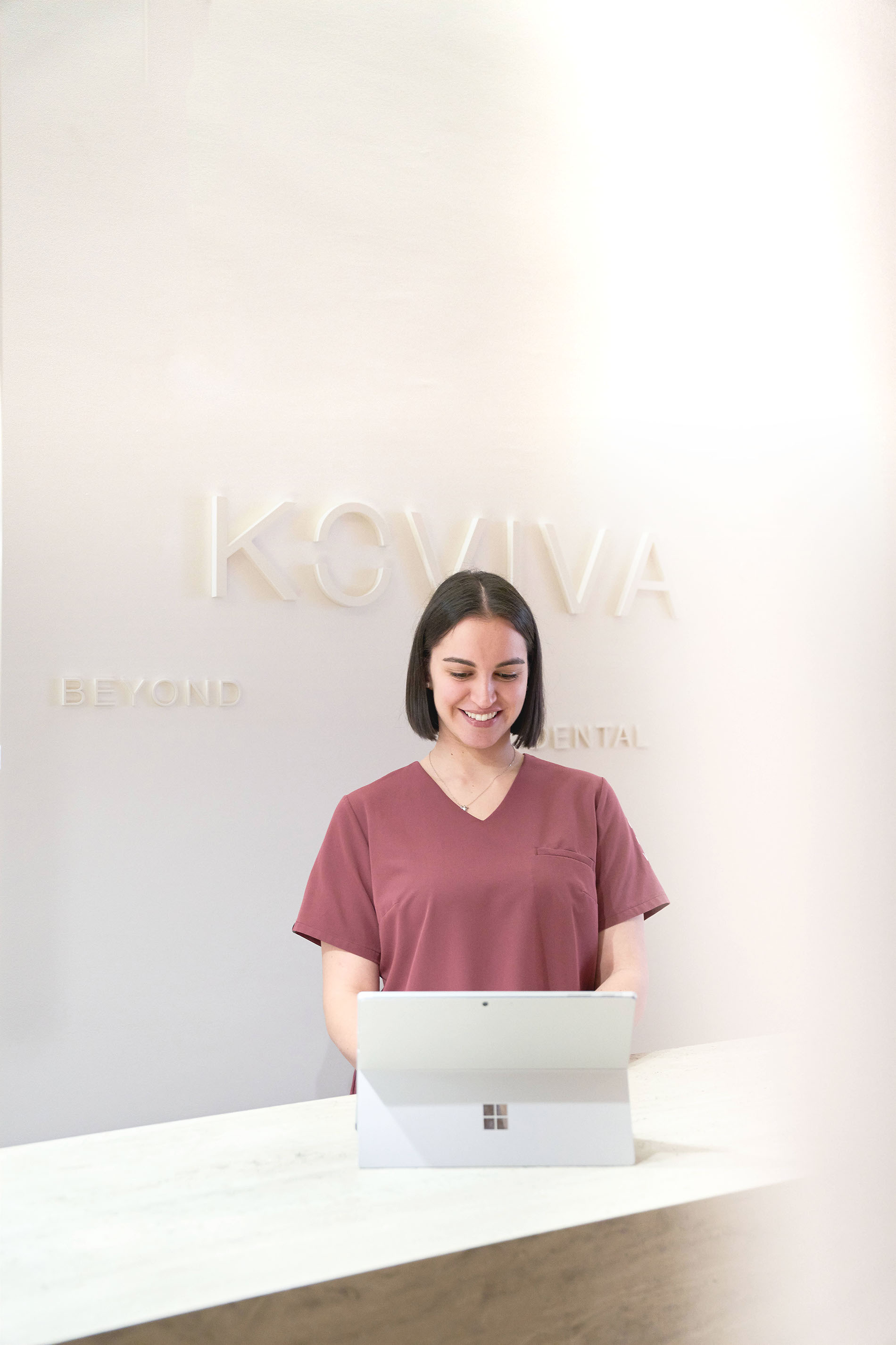 Lächelnde Frau in medizinischer Kleidung am Empfangstresen mit Laptop, Praxis KOVIVA DENTAL