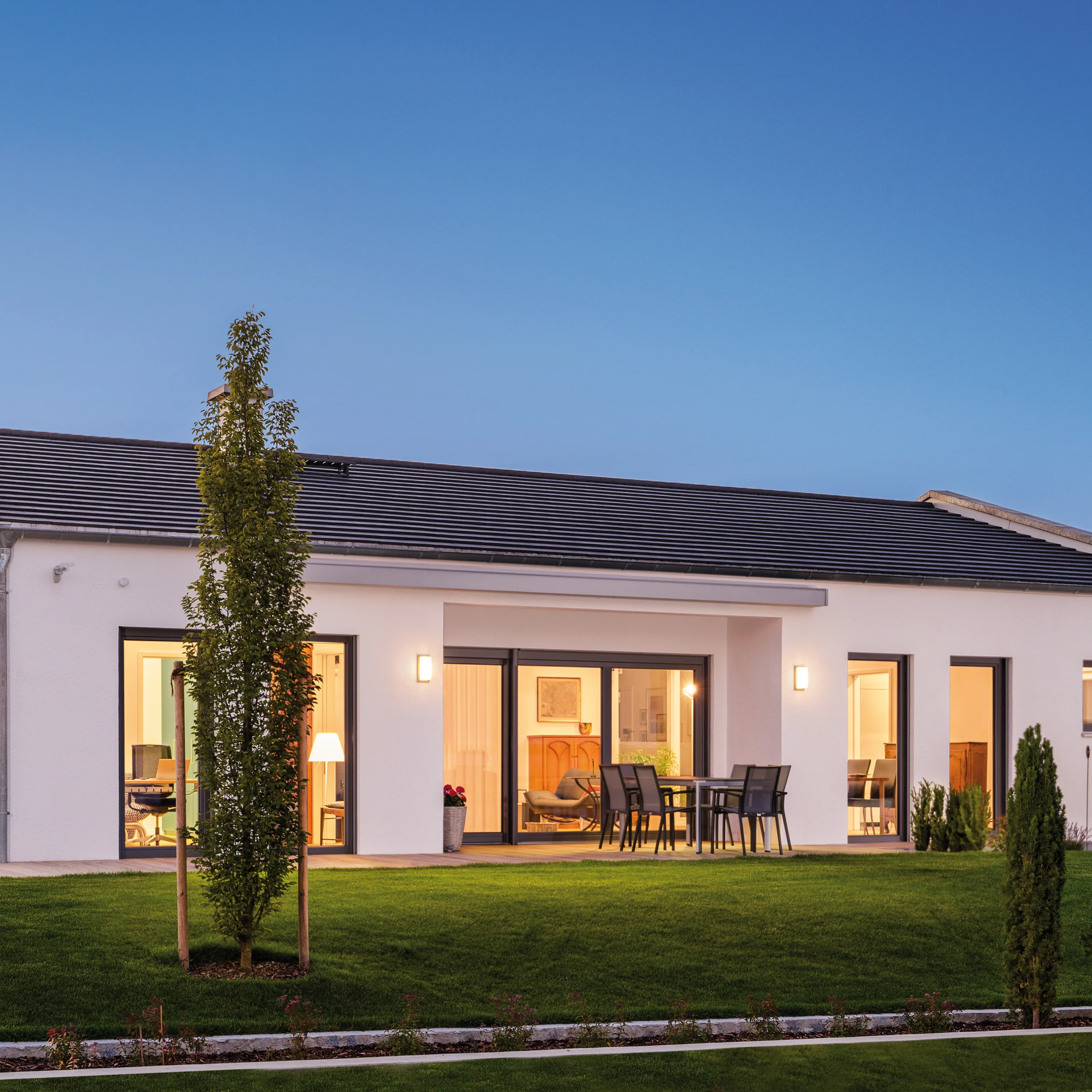 Modernes weißes Einfamilienhaus mit großen Fenstern und Terrasse, beleuchteter Garten im Abendlicht.