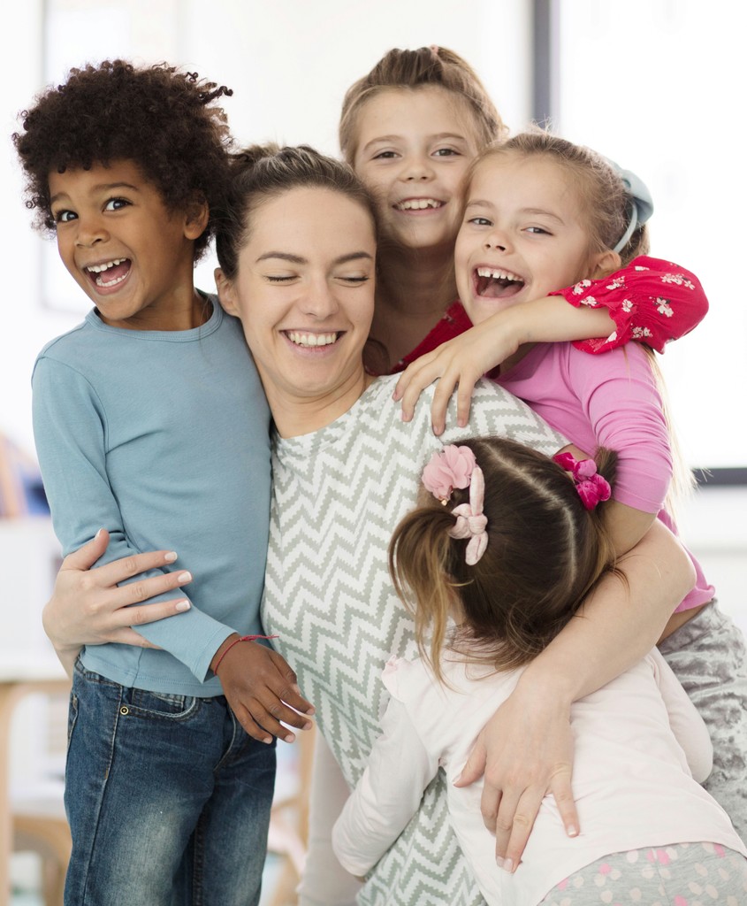 A smiling woman enjoying a joyful group hug with four cheerful children in a bright and happy setting, showcasing warmth and affection.