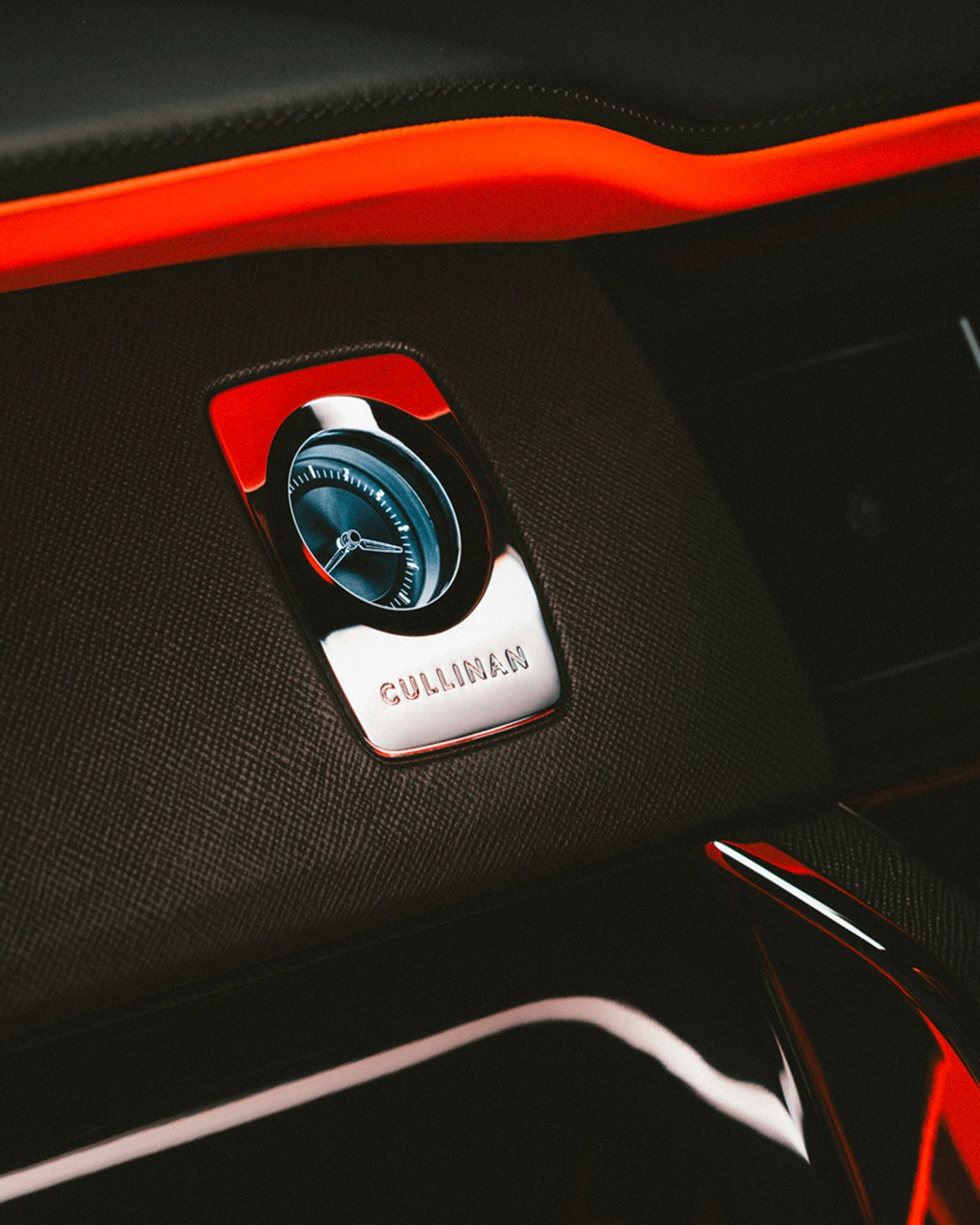 Interior detail of a Rolls-Royce Cullinan showing the chrome-framed analogue dashboard clock with "CULLINAN" engraved beneath it, set into black leather with orange ambient trim.