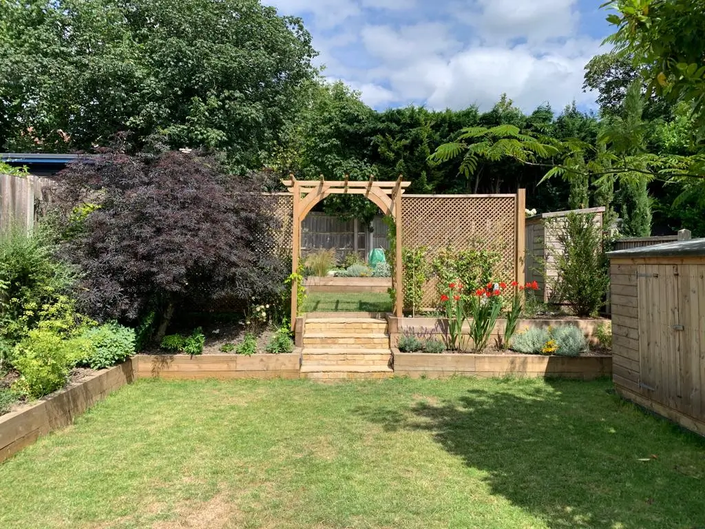 A garden scene featuring a stone archway, lush plants, and a manicured lawn under a partly cloudy sky.