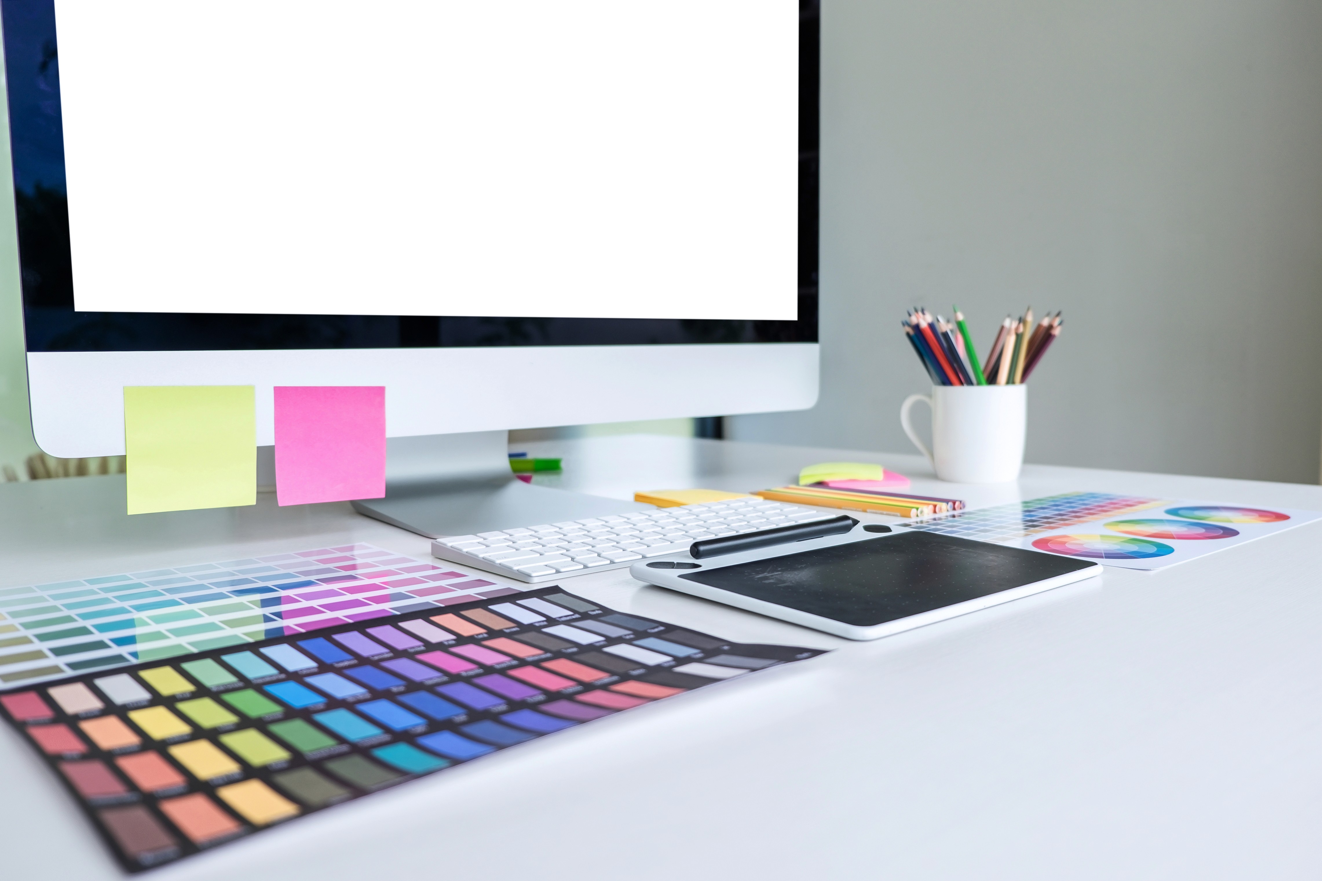 Graphic designer workspace with digital drawing tablet, color palettes, sticky notes, and a computer on a clean white desk.